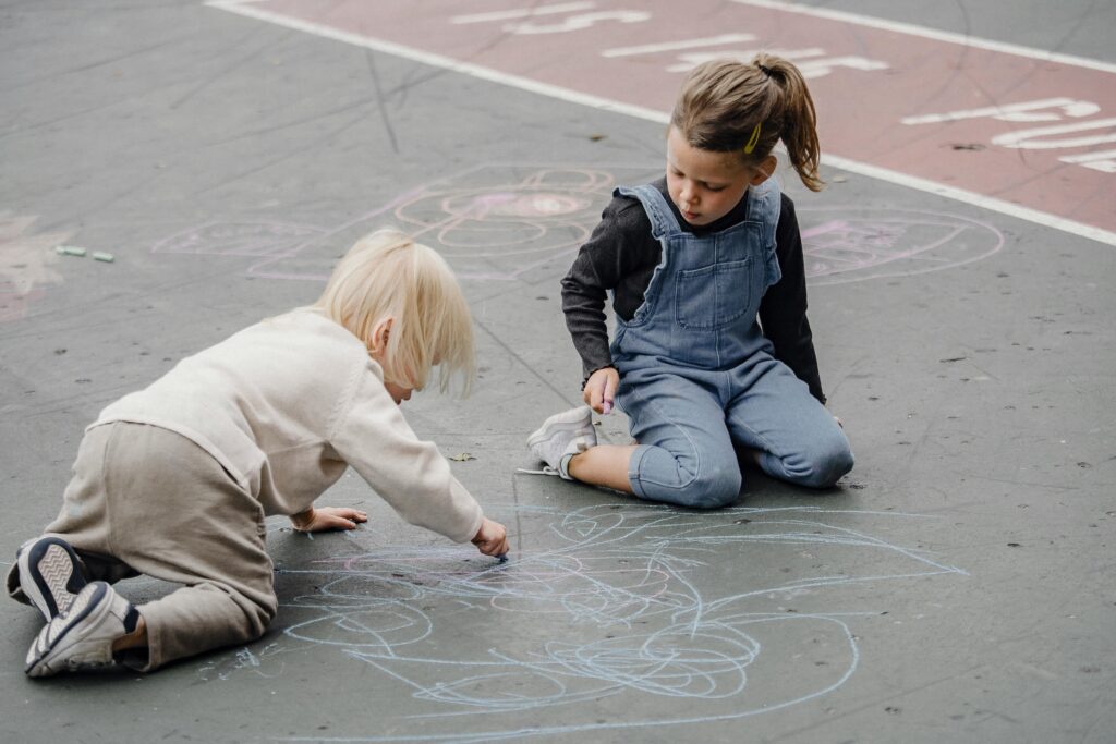Two children play on the concrete outdoors as they draw with colored sidewalk chalk.
